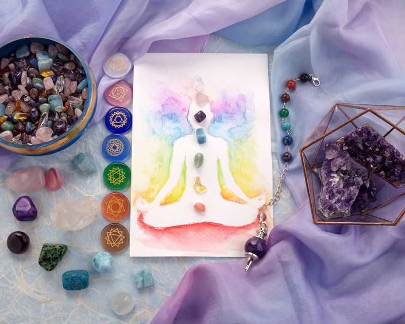 A drawing of a person meditating is covered with gemstones in the 7 chakra colors. chakra symbols, small gemstones, and an amethyst sit by it on a table.