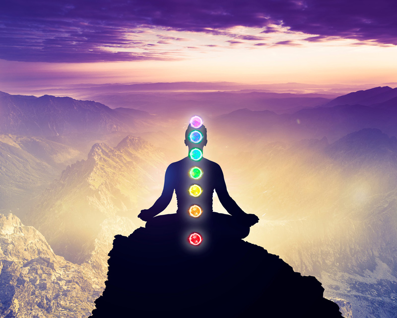 A man is meditating in a yoga pose while sitting on a mountain high in the sky with the 7 colors of the chakras displayed over his body.