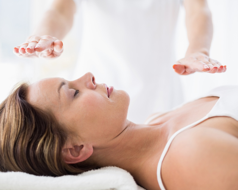 A woman lies peacefully on her back while a Reiki Healer's hands are held steadily above her body to perform Reiki Healing.